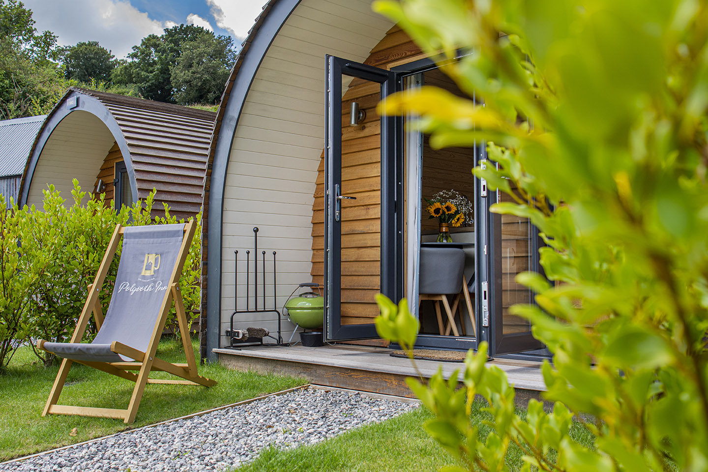 Image shows the front view of the garden glamping pods with greenery around it, and a deckchair on the lawn