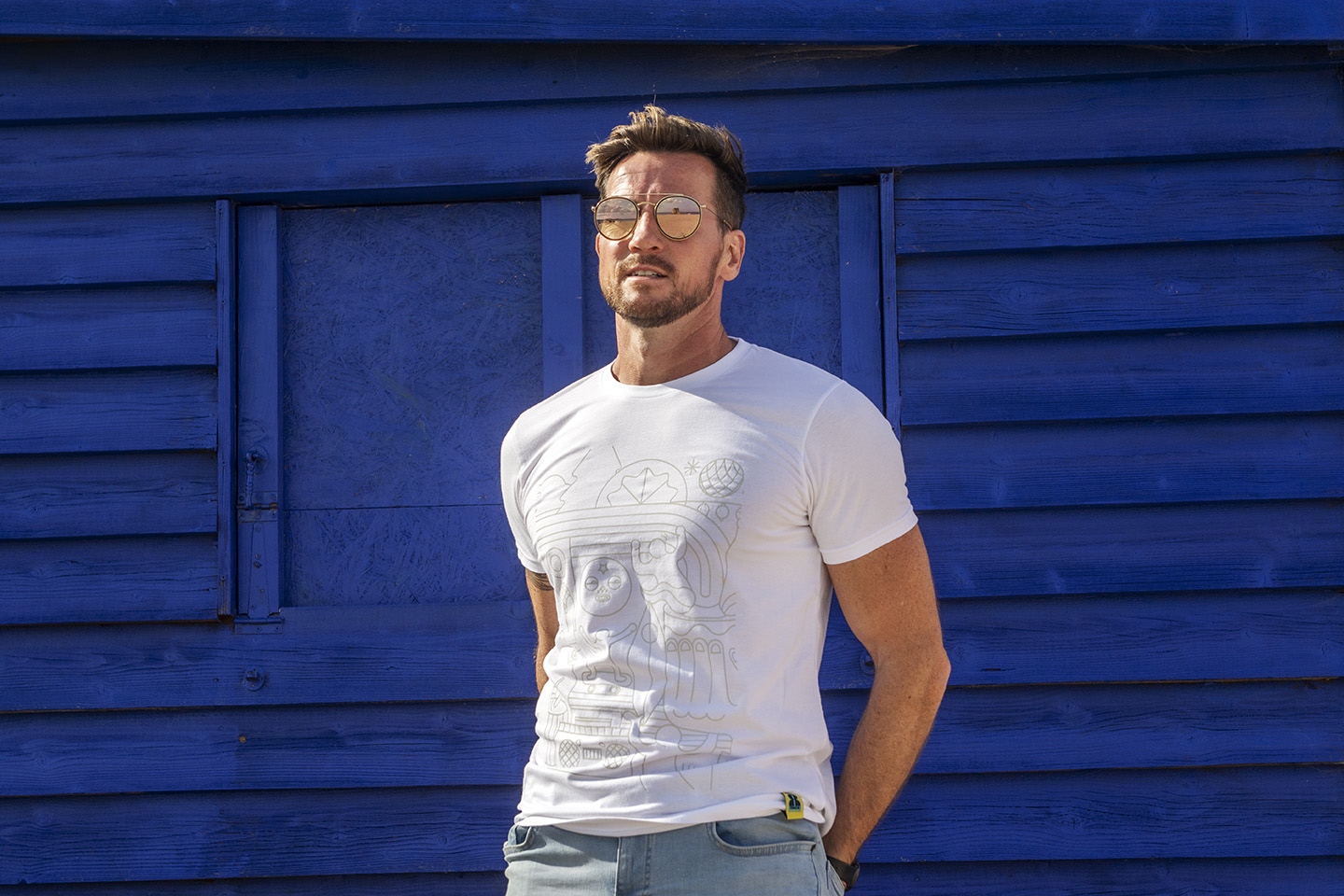 A male model stands in front of rich royal blue painted wooden background, wearing a white 8Track Rum branded t-shirt and mirrored sunglasses.