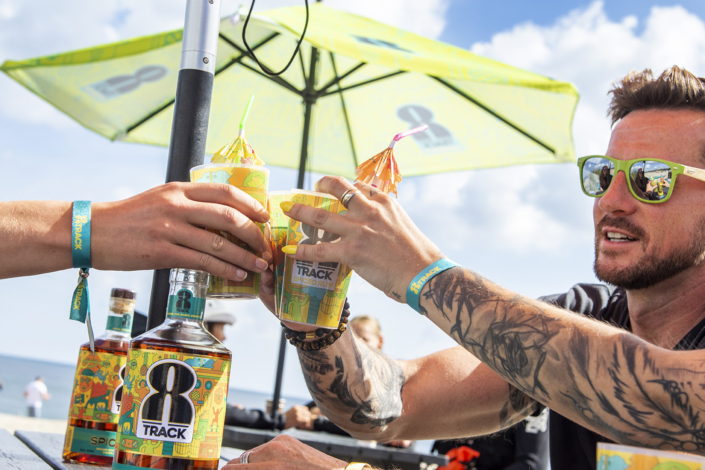 Three models holding up cups of 8Track Rum in a cheers pose, in an outdoor bar setting on a beach with umbrellas in the background