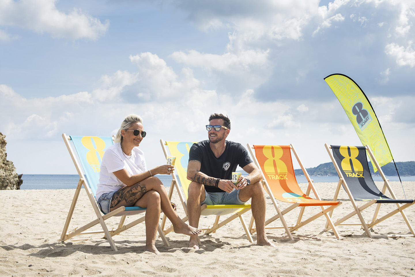 A male and female model sit together on 8Track Rum branded deckchairs in the sunshine on a beach in Cornwall. Blue sky and clouds overhead.