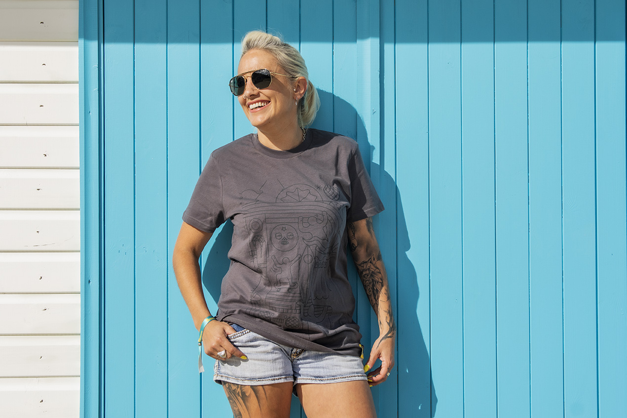 A female model stands in the sunshine against a white and turquoise wooden background wearing a dark grey 8Track branded t-shirt 