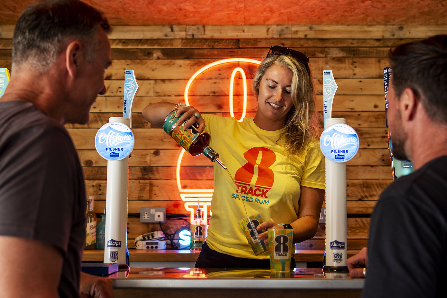 A female bar tender pours 8Track rum from a height into a glass at a bar with two male models looking on. In the background is a neon 8Track logo sign against a timber background.