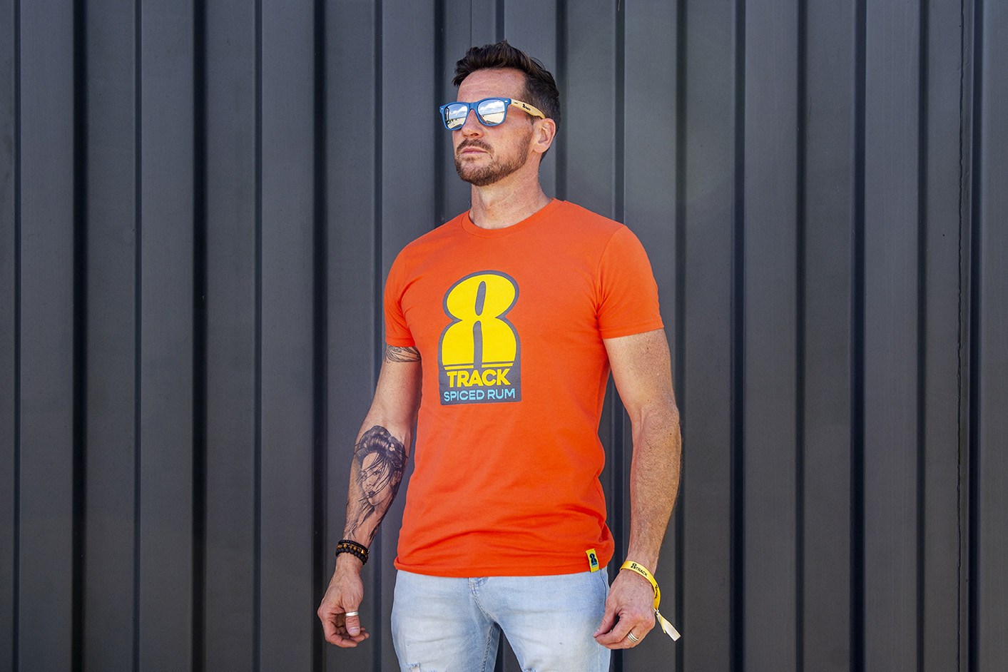 A male model stands in front of a dark grey panelled background in an orange 8Track t-shirt and 8Track mirrored sunglasses