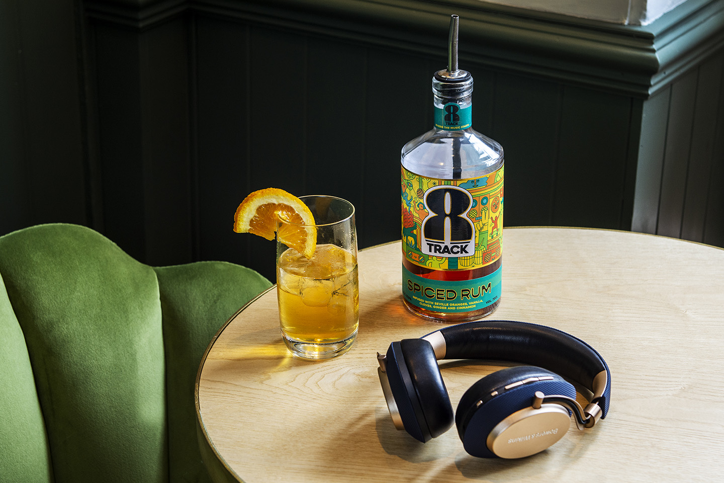 Bottle of 8Track Rum on a table with a glass of rum over ice with a slice of orange on the side. A pair of headphones sit on the table as well with a green chair to the left.