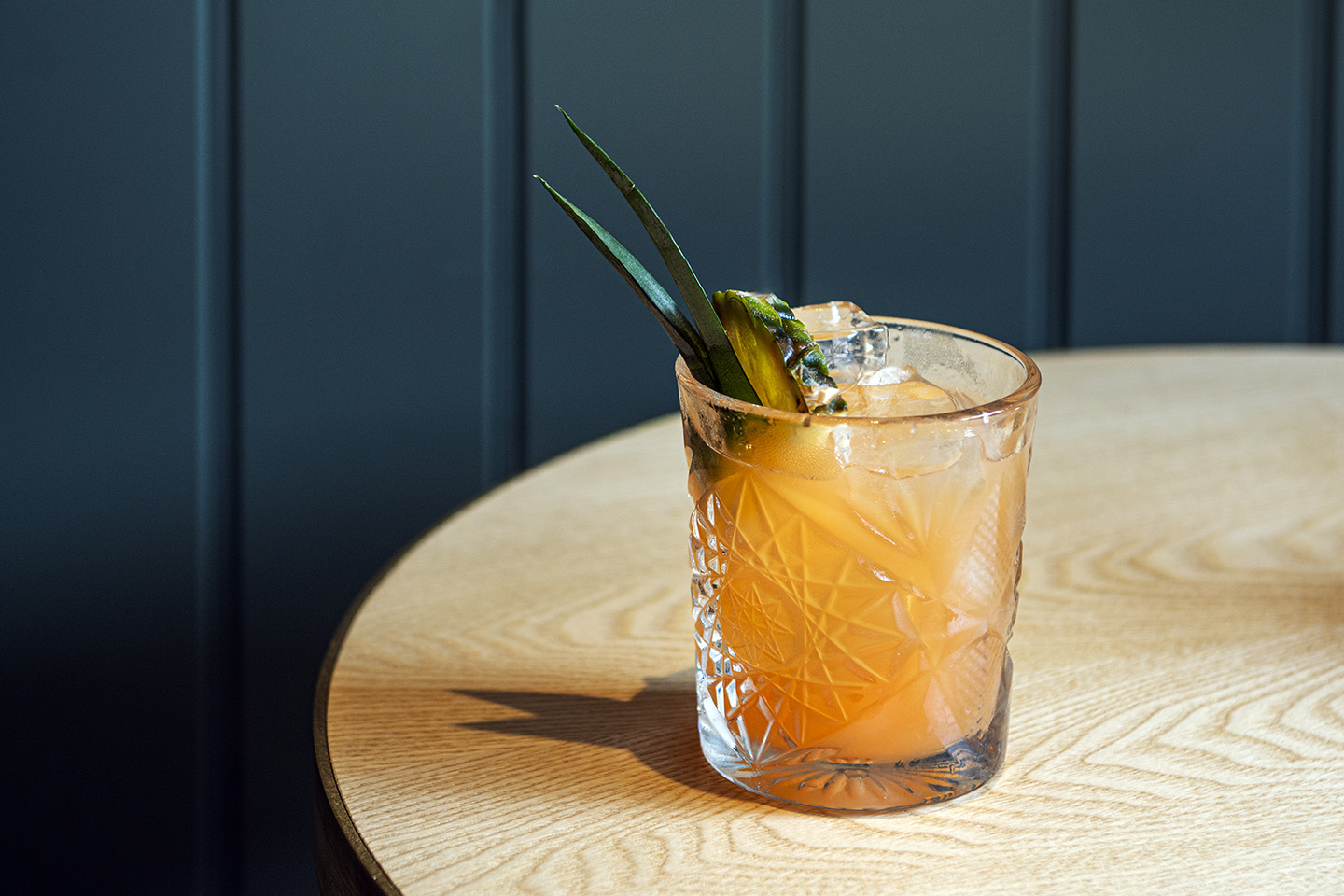 Beautiful etched glass with the orange coloured 'Welcome to The Jungle' cocktail in it with a pineapple leaf and slice garnish, on a round table with dark painted wood background.