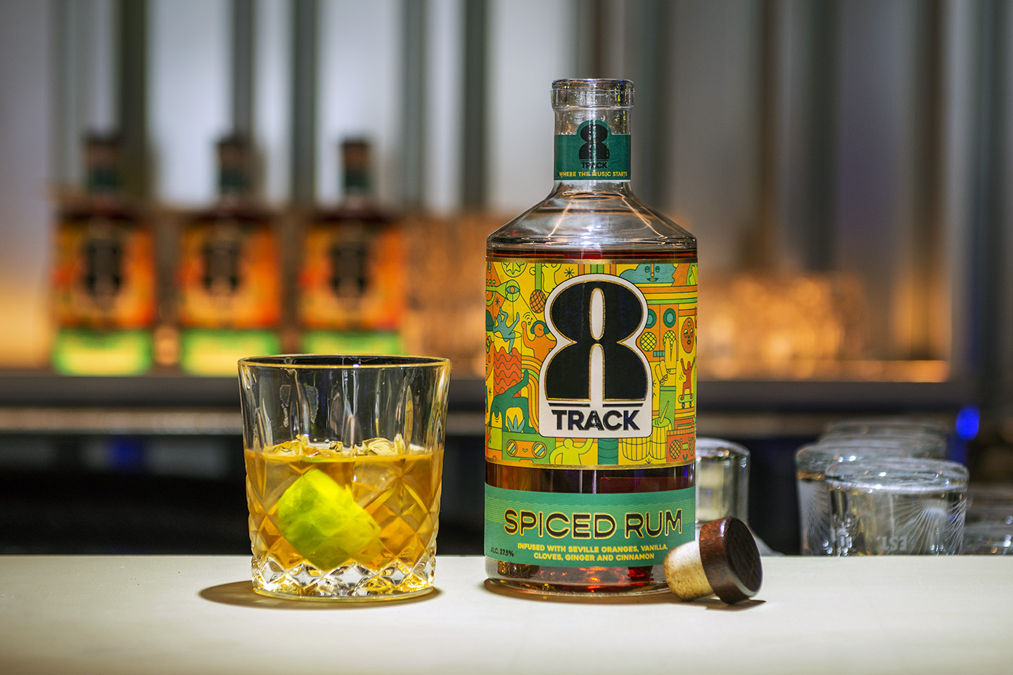 A bottle of 8Track Rum on a white top bar with glass of rum over ice with a wedge of lime. Bottles of rum are lined up in the background with atmospheric lighting and corrugated metal backdrop