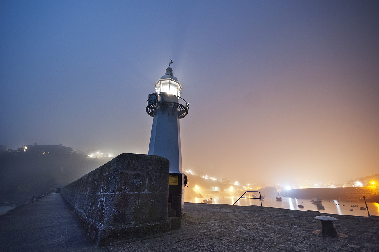 View of the lighthouse shining at Mevagissey harbour on a misty dark evening.