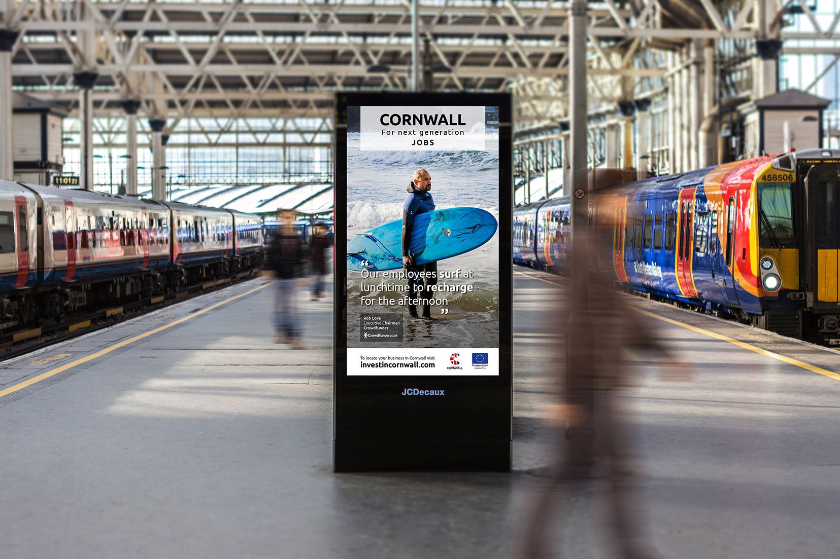 A JC Decaux digital billboard at a busy London station with a portrait of Rob Love holding a surfboard on it