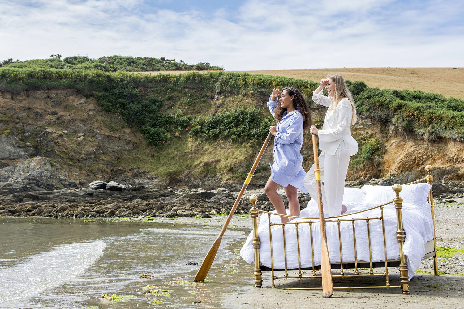 Two female models dressed in pyjamas stand on a large brass bed on a beach whilst holding boat oars and looking out to sea whilst the tide laps at the bed