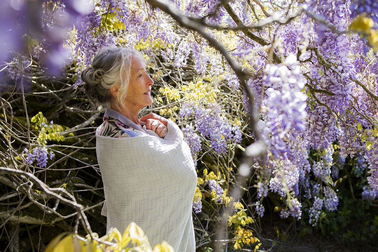 model wraps herself in a throw in the grounds of Caerhays Castle surrounded by lots of beautiful purple wisteria in the background