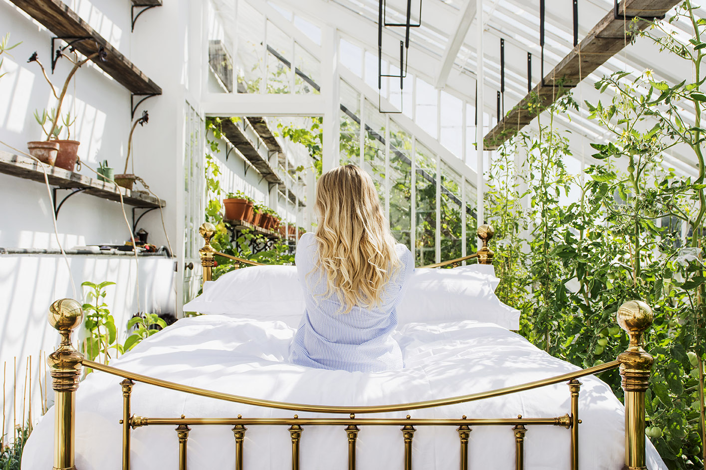 A female model sits with her back to the camera on a large brass bed with crisp white linens in the old glasshouse at the Lost Gardens of Heligan