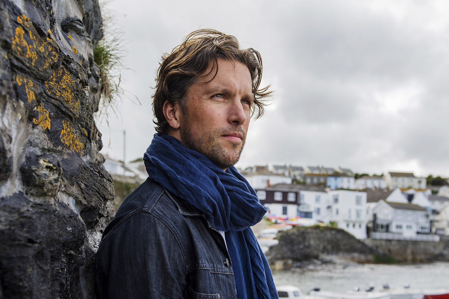 a male model with a dark blue linen scarf on looks out to sea with a fishing village in the background