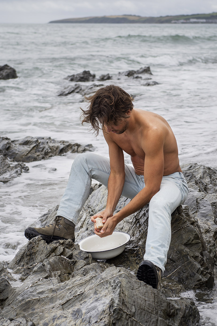 a male model sits on a rock with a tin bowl using seaweed soap to wash with the ocean in the background