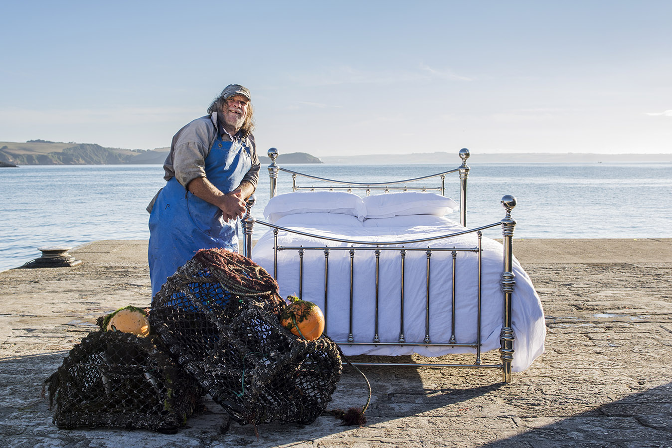 a salty sea dog fisherman called Malcolm stands next to a brass bed with crisp white linens on the quay at Mevagissey Harbour. There are lobster pots in the foreground.