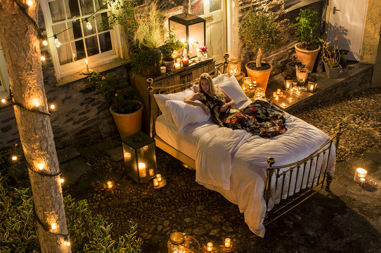 a female model reclines on a large brass bed in an enclosed courtyard at night surrounded by lit candles and lanterns