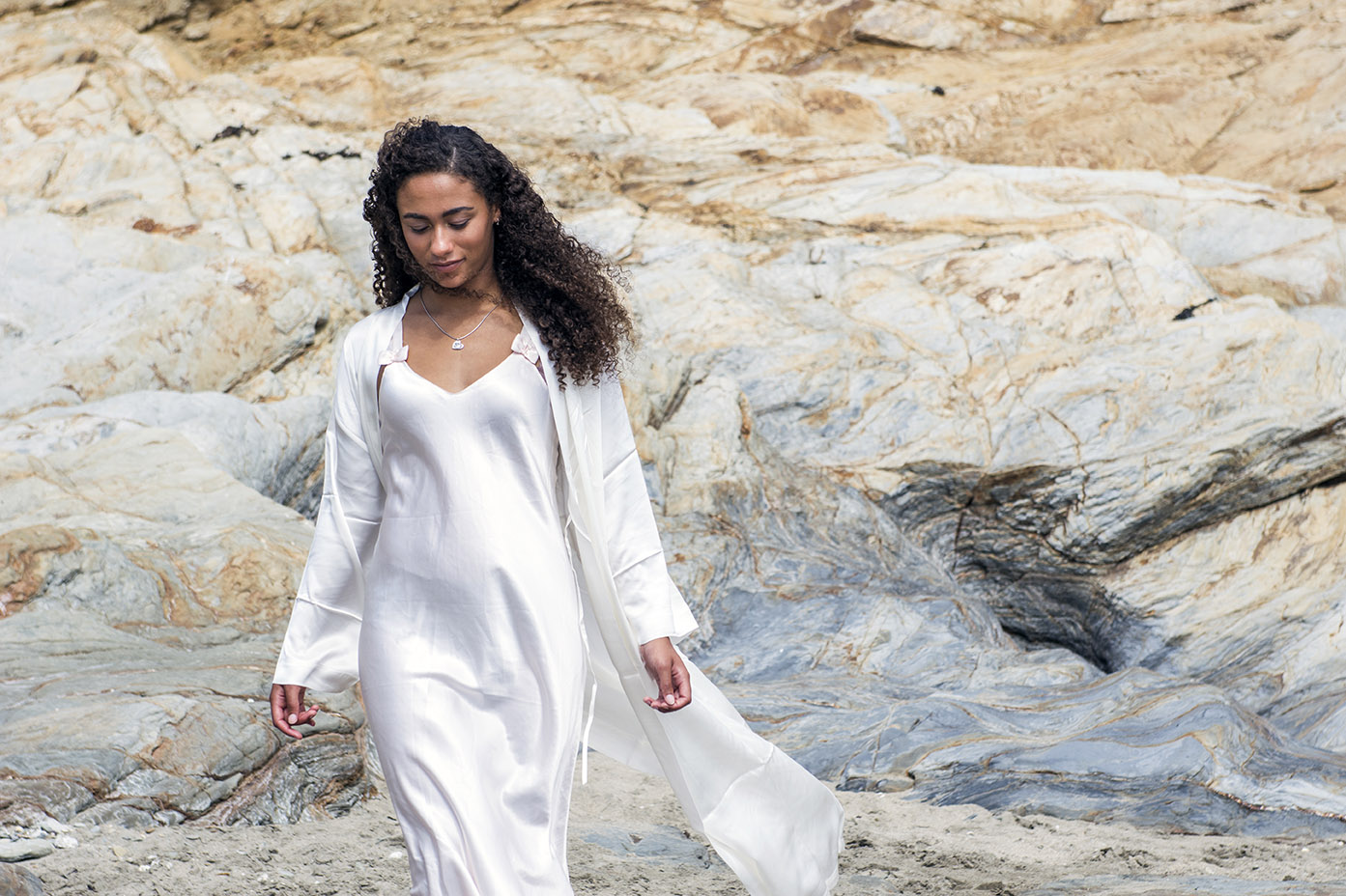 a female model dressed in white silk pyjamas walks on the beach with sandy coloured rocks in the background