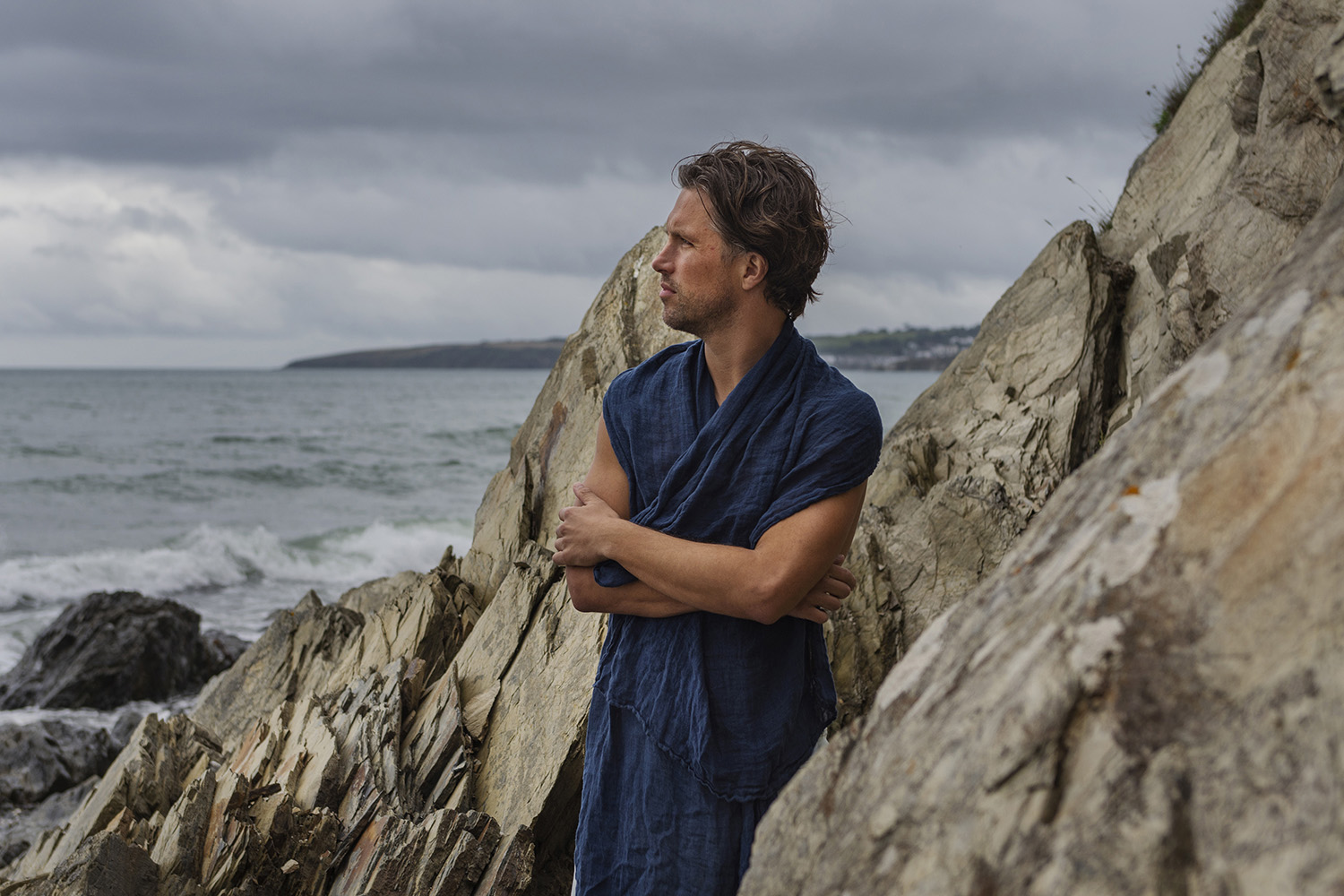 a male model gazes out to sea, wrapped in a dark blue linen scarf with rocks and coastline in the background 
