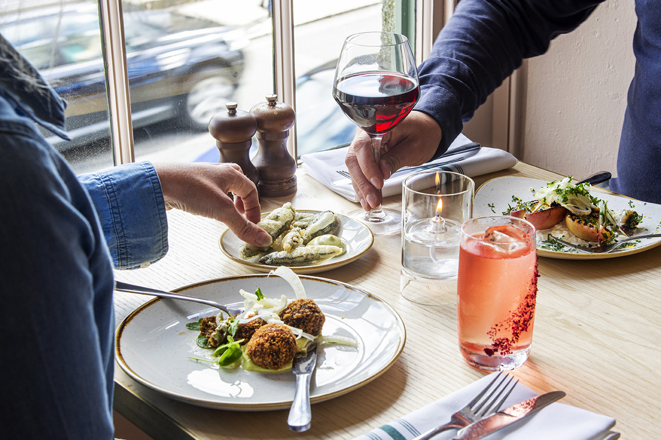 A couple dining in the window of Lemon Street Longstore with several small plates of food a glass of red wine and a cocktail