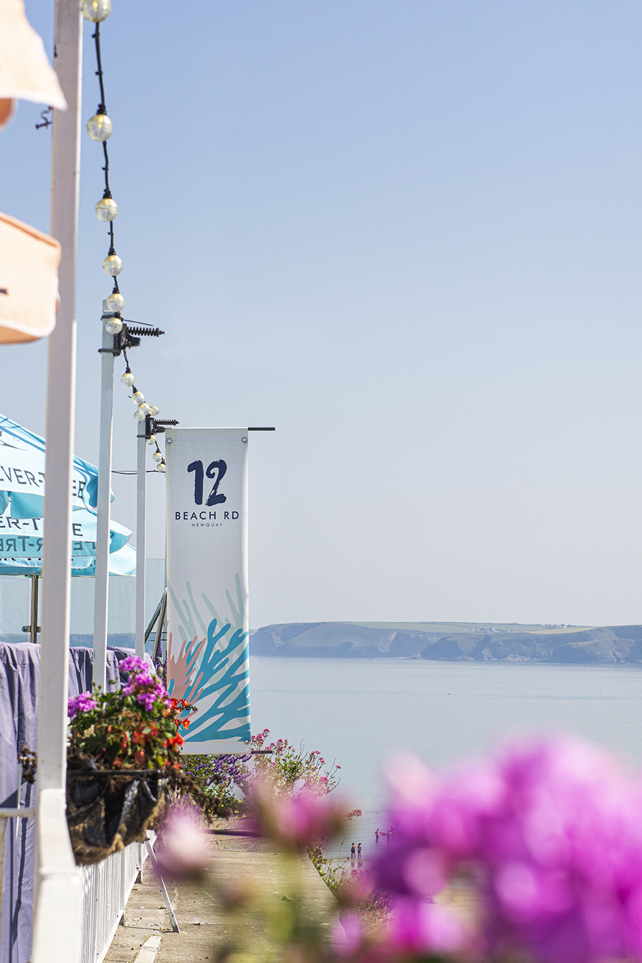 Exterior view of 12 Beach Road at Newquay on a bright summers day, pink flowers in the foreground and the sea and coastline in the distance