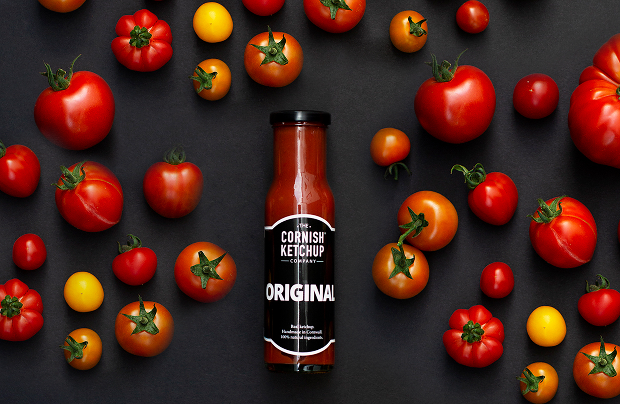 A bottle of Cornish Ketchup on black background surrounded by all sorts of different types of tomatoes