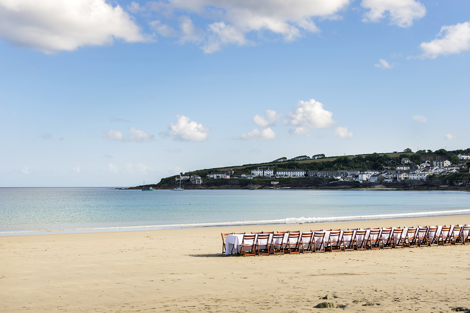 View of an empty Porthcurnick beach with a long table set for dinner, the village of Portscatho is in the background