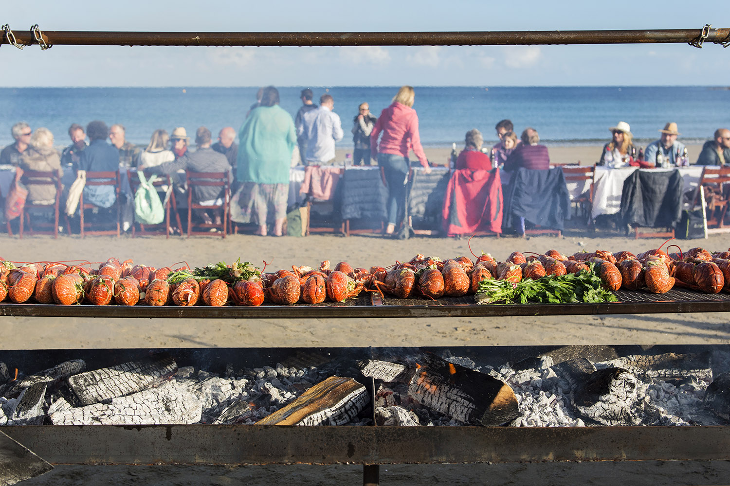 View of large wood fired grill on Porthcurnick Beach with lobsters cooking in the foreground and diners at a long table in the background